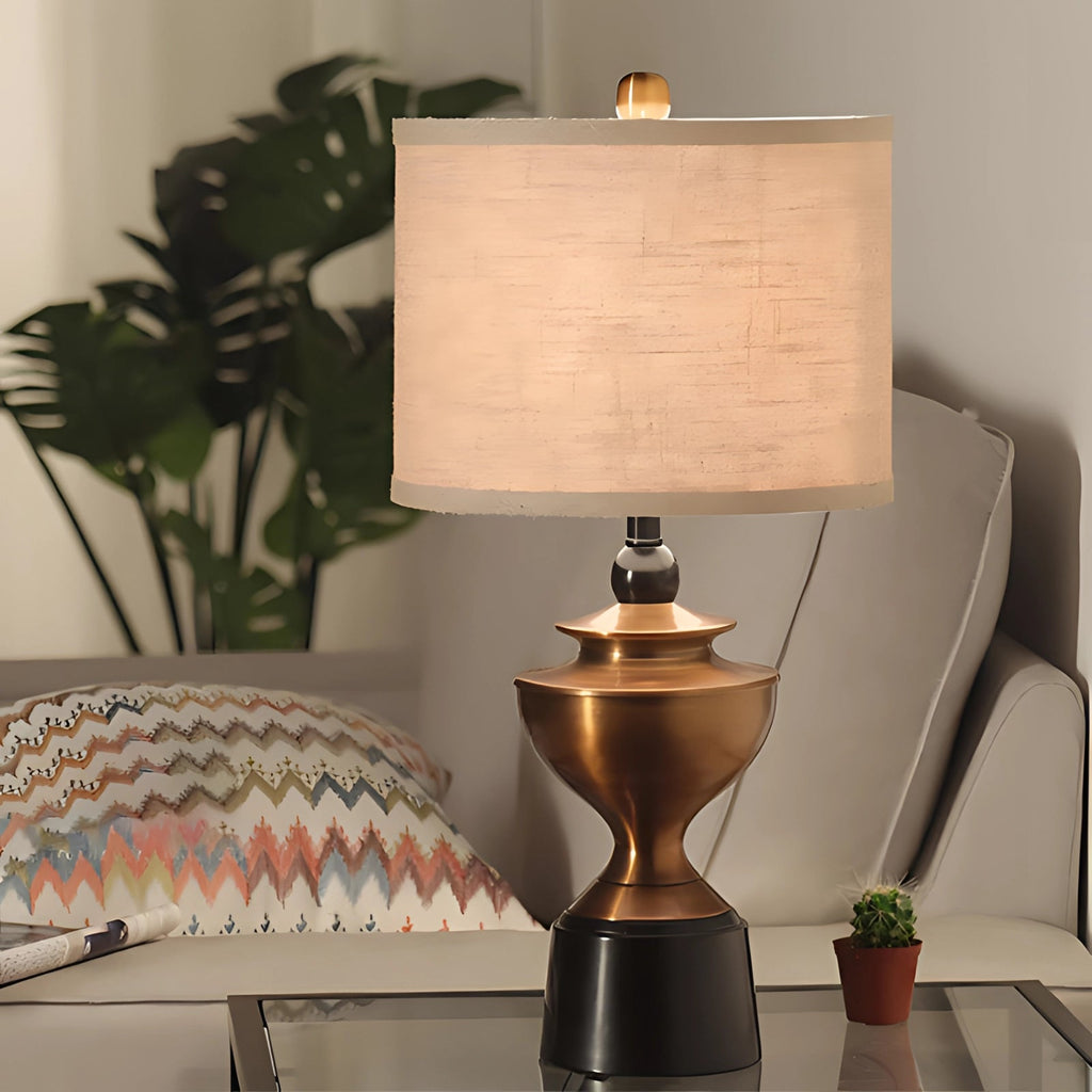 Bronze table lamp with a square shade, situated on a wooden table, accompanied by houseplants and a flowerpot, enhancing the elegant interior design of a living room setting.